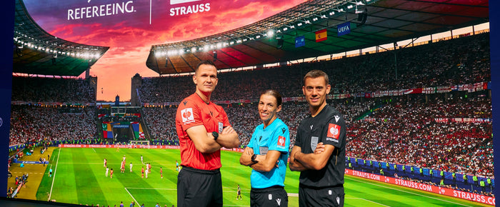 Drei UEFA-Match-Officials stehen vor einer Stadionkulisse. Auf den Ärmeln ihrer roten, blauen und schwarzen Trikots ist jeweils das STRAUSS-Logo sichtbar.