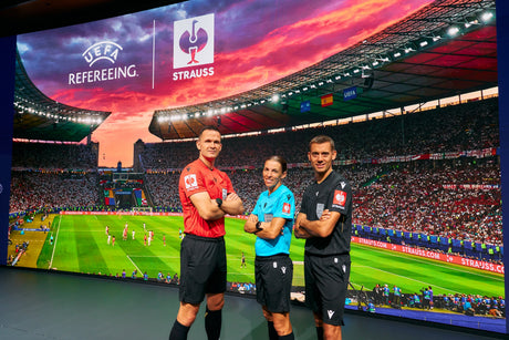 Drei UEFA-Match-Officials stehen vor einer Stadionkulisse. Auf den Ärmeln ihrer roten, blauen und schwarzen Trikots ist jeweils das STRAUSS-Logo sichtbar.