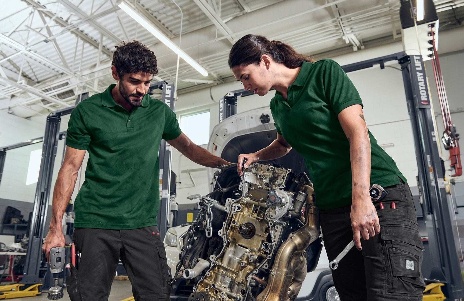 Mann und Frau bei der Reparatur eines Motorblocks in grünen Arbeitsshirts und schwarzen Arbeitshosen. 