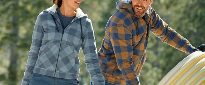 Eine Dame und ein Herr beim Arbeiten im Winter - die Sonne scheint, Schnee liegt. Sie tragen beide die Karo-Kapuzenjacke e.s.iconic: Flanell mit durchgehendem Faserpelz-Futter